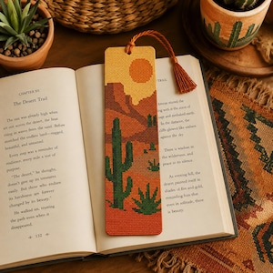 May include: A rectangular cross-stitch bookmark featuring a desert landscape with a yellow sun, brown rock formations, and green cacti. The bookmark has a textured grid pattern and is finished with a matching orange tassel attached to the top.