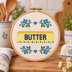 May include: A circular wooden embroidery hoop containing a cross-stitch design on white fabric. The design features a yellow stick of butter with the word BUTTER in blue text, surrounded by blue flowers with green leaves and small blue hearts.