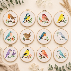Op de afbeelding: Twaalf ronde houten borduurringen met kruissteekpatronen van diverse vogels op takken. Elke ring toont een unieke vogelsoort in levendige kleuren, waaronder blauw, geel, rood en bruin, op een witte stoffen ondergrond.