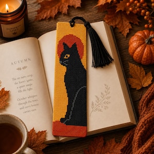 May include: A rectangular cross-stitch bookmark featuring a black cat silhouette against a yellow background with a red circular sun motif. The bookmark is finished with a black thread tassel attached to the top and has a textured grid pattern.