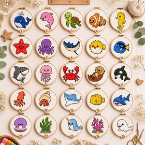 May include: Twenty-five circular wooden embroidery hoops containing cross-stitch patterns of various sea creatures, each numbered one through twenty-five. The designs include a whale, dolphin, turtle, fish, seahorse, starfish, octopus, ray, pufferfish, shark, jellyfish, crab, otter, orca, squid, hermit crab, stingray, boxfish, whale shark, clam, seaweed, narwhal, coral, and beluga.
