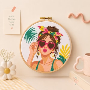 May include: A cross-stitch embroidery in a round wooden hoop featuring a person with brown hair wearing a colorful floral headscarf and pink heart-shaped sunglasses. The design includes green palm leaves and yellow foliage on a white fabric background.