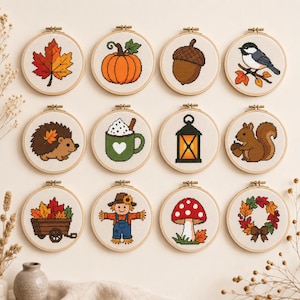 Può includere: Dodici telai da ricamo circolari con disegni a punto croce a tema autunnale, tra cui una foglia d'acero, una zucca, una ghianda, un uccellino, un riccio, una tazza, una lanterna, uno scoiattolo, un carro, uno spaventapasseri, un fungo e una ghirlanda su tessuto bianco.
