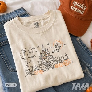 Op de afbeelding: Een ivoorkleurig t-shirt met een zwart-oranje lijntekening van een spookkasteel, een heks op een bezemsteel, vleermuizen, een kat en pompoenen. Het shirt heeft een Comfort Colors-label in de nek en bevat een afbeelding van een hek.