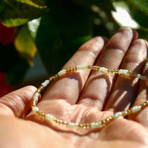 May include: A delicate beaded bracelet with gold and pale green beads. The bracelet is held in an open hand, with sunlight illuminating the details. The bracelet's design features small, evenly spaced beads, creating a subtle and elegant accessory.