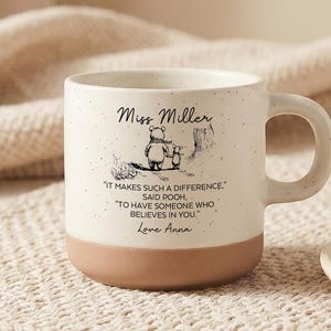 Könnte beinhalten: Cremefarbene Keramik-Tasse mit braunem Boden und Henkel. Die Tasse zeigt eine Winnie-Puuh-Illustration und den Text "Miss Miller" und ein Zitat. Ein kleiner Teller mit einem Keks befindet sich im Hintergrund.