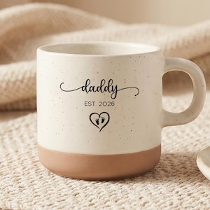 Puede incluir: Taza de cerámica moteada con base marrón y asa curva. La taza presenta la palabra "daddy" en fuente cursiva, el año "EST. 2026" y un corazón con huellas. Una galleta está sobre un plato pequeño.