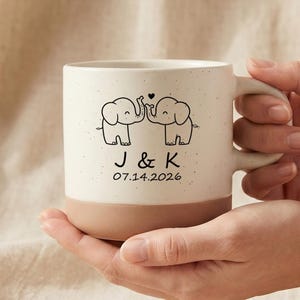 以下が含まれることがあります： クリーム色の斑点模様の上部と無地のベージュ色の底部を持つ、ツートンカラーのセラミック製マグカップ。前面には、2頭の象と小さなハート、アルファベットのJとK、そして2026年7月14日の日付が黒いインクで描かれています。