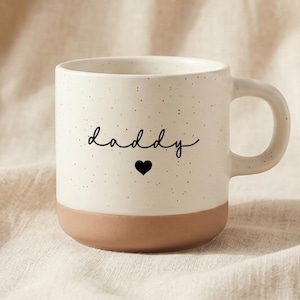 Puede incluir: Taza de cerámica color crema con acabado moteado y base marrón. La palabra "daddy" está escrita en cursiva, con un corazón negro debajo. La taza tiene un asa curvada.