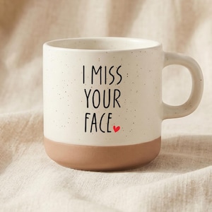 Puede incluir: Una taza de cerámica con una sección superior de color crema moteado y una base de color terracota sólido. El texto "I MISS YOUR FACE" está impreso en letras negras sin remates en el frente, acompañado por un pequeño icono de corazón rojo.