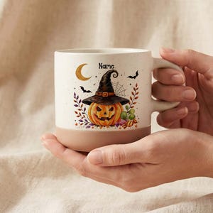 May include: A cream-colored ceramic mug with a two-tone design featuring a tan base. The front displays a watercolor illustration of a jack-o'-lantern wearing a witch hat, a crescent moon, bats, and colorful autumn leaves with the text Name.