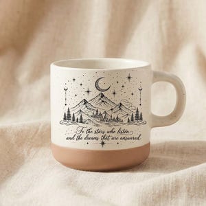 May include: A ceramic mug with a speckled cream upper section and a solid terracotta base. The front features a black line art illustration of mountains, pine trees, a crescent moon, and stars, with the text: To the stars who listen and the dreams that are answered.