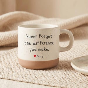 Könnte beinhalten: Ein gesprenkelter, cremeweißer Keramikbecher mit einem soliden, terrakottafarbenen Boden. Auf der Vorderseite steht der schwarze Text "Never forget the difference you make.", gefolgt von einem kleinen roten Herz und dem Namen "Daisy".