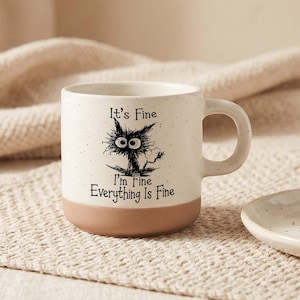 May include: A speckled off-white ceramic mug with a brown base, featuring a black ink illustration of a wide-eyed, dishevelled cat. The text printed on the front reads: It's Fine, I'm Fine, Everything Is Fine, in a simple black sans-serif font.