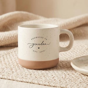 Könnte beinhalten: Ein cremefarbener Keramikbecher mit gesprenkelter Oberfläche und einem terrakottafarbenen Boden. Auf der Vorderseite steht in schwarzer Schrift Promoted to grandma Est. 2026 mit einem kleinen Herzsymbol. Der Becher hat einen runden Henkel.