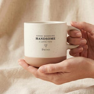 May include: A ceramic mug with a speckled cream upper section and a solid terracotta base. The front features the black printed text GOOD MORNING HANDSOME I LOVE YOU, a small hand-drawn heart, and the name Daisy centered below the heart.