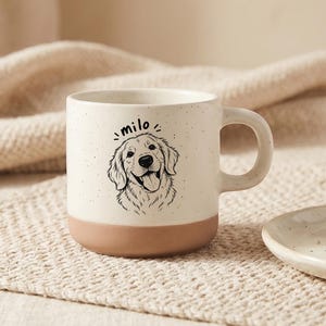 May include: A cream-colored ceramic mug with a speckled finish and a solid terracotta-colored base. The front features a black line-art illustration of a golden retriever dog with the name "milo" written in lowercase script above the dog's head.