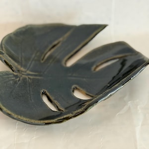 May include: A dark green ceramic dish shaped like a monstera leaf. The surface has a glossy finish with visible veins and cutouts characteristic of the plant. The edges are slightly curved upward, creating a shallow basin for holding small items.