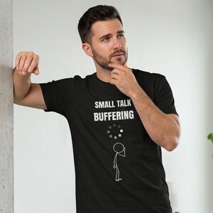 May include: Black t-shirt with the text "SMALL TALK BUFFERING" in white, along with a graphic of a stick figure and a loading symbol. The person is wearing dark blue jeans and white sneakers.