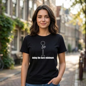 Puede incluir: Camiseta negra con un diseño de figura de palo blanco y la frase "doing the bare minimum". La camiseta es llevada por una persona frente a un fondo borroso al aire libre.