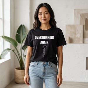 Pode incluir: T-shirt preto com o texto branco "OVERTHINKING AGAIN" e um desenho de boneco palito. A t-shirt é usada com calças de ganga azuis. A imagem mostra uma mulher a usar a t-shirt numa divisão com uma planta grande no fundo.