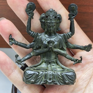 Puede incluir: Estatua de color bronce de una deidad sentada con múltiples brazos, sosteniendo varios objetos. La figura tiene un tocado adornado y está en una pose meditativa. La estatua es detallada y parece ser de origen asiático.