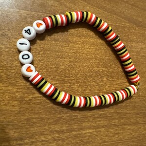 May include: A beaded bracelet with the word "4100" spelled out in white letter beads. The bracelet features alternating red, white, black, and yellow disc beads. The bracelet is on a wooden surface.