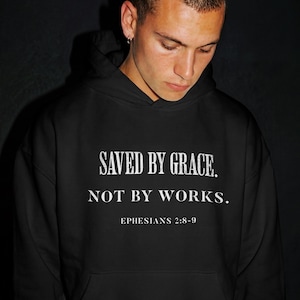 Könnte beinhalten: Schwarzer Kapuzenpullover mit weißem Text: "SAVED BY GRACE. NOT BY WORKS. EPHESIANS 2:8-9." Der Hoodie hat eine Kapuze mit Kordelzug und eine Kängurutasche. Die Person, die den Hoodie trägt, schaut nach unten.