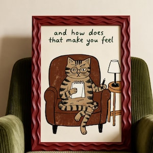 May include: A framed print in a wavy dark red frame featuring a cartoon illustration of a tabby cat wearing glasses, sitting in a brown armchair, holding a notepad and pen, with the text "and how does that make you feel" written above.