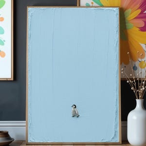 May include: A rectangular framed wall art piece featuring a textured light blue painted background with a small, detailed illustration of a penguin chick standing in the lower center. The frame is a simple, thin, light-colored wood finish.