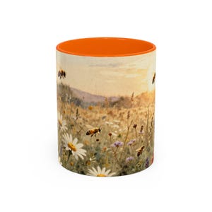 Könnte beinhalten: Ein Keramikbecher mit leuchtend orangefarbenem Innenraum und Rand. Die Außenseite zeigt eine aufgedruckte Illustration einer sonnendurchfluteten Wiese mit weißen Gänseblümchen, Wildblumen und mehreren Bienen, die zwischen den Pflanzen fliegen.