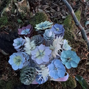 May include: A decorative bouquet composed of various pine cones painted in shades of blue, purple, and green, interspersed with white artificial foliage. The arrangement features multiple flower-shaped pine cone heads and a light pink ribbon base.