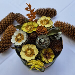 Op de afbeelding: Een ronde houder gevuld met een decoratief arrangement van dennenappels en handgemaakte bloemen van dennenappelschubben. De bloemen zijn geschilderd in tinten geel, wit en groen, geaccentueerd door bruine dennenappels en een gedroogde oranje tak.