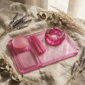 May include: A rectangular pink translucent rolling tray featuring a circular grinder, a central rectangular slot, and a matching pink ashtray. The tray is constructed from a smooth, clear material with a vibrant magenta hue and defined edges.