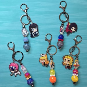 May include: Five metal keychains featuring acrylic anime character charms and colorful beaded accents. Each keychain includes a lobster clasp, a split ring, and a dangling charm of a character paired with spherical beads in various colors.