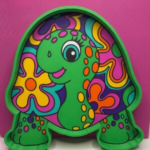 May include: A green turtle-shaped wall art piece featuring a cartoon turtle with a bright green head and neck, decorated with orange and yellow spots. The shell displays a colourful pattern of yellow, orange, and purple flowers.