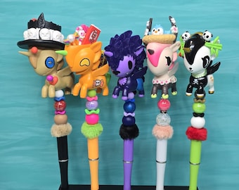 May include: Five decorative pens featuring colorful creature toppers, including a skull-adorned tan figure, an orange character, a purple creature, a pink-eared animal, and a black-and-white panda-themed figure, all displayed in a black stand labeled GEEKSPOOL.