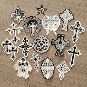 Peut inclure: Une collection d'autocollants noirs et blancs représentant divers motifs de croix, une fleur de lys, une étoile et un emblème circulaire. Un autocollant circulaire porte le texte Chrome Hearts. Les motifs incluent des filigranes et des ailes.