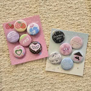 May include: Two sets of circular pin-back buttons on display cards. The pink card features six buttons with strawberry, floral, heart, and pixelated designs. The light blue card features five buttons with star, music note, and house graphics with text.