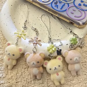 Puede incluir: Funda de teléfono transparente con diseño de caras sonrientes y colgantes de ositos de peluche. Los colgantes son de varios colores, incluyendo amarillo, rosa y beige. La funda tiene la frase "Be happy" impresa.