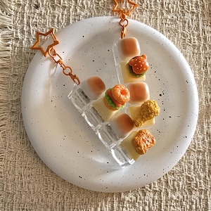 May include: A clear acrylic letter V keyring featuring miniature food charms including small bread rolls, a burger, a doughnut, and fried chicken pieces, attached to an orange star-shaped metal clasp and a chain link connector.