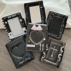 Peut inclure: Plusieurs albums photo noirs et blancs avec divers motifs. Certains albums présentent des formes de cœur, des illustrations de chats et du texte tel que "Angel Bunny" et "Exchange". Les albums sont disposés sur une surface en bois.