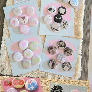 Könnte beinhalten: Eine Sammlung runder Ansteckbuttons, die auf acht rechteckigen Präsentationskarten angeordnet sind. Die Buttons zeigen verschiedene grafische Motive wie Erdbeeren, Herzen, Noten, Sterne, Flügel und diverse Schriftzüge in verschiedenen Farben.