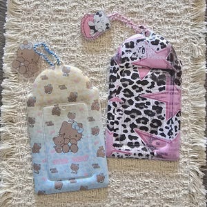 Puede incluir: Dos tarjeteros rectangulares con parte superior redondeada. El tarjetero izquierdo es azul claro y amarillo con un patrón de Hello Kitty y ositos de peluche y una cadena de cuentas azul. El derecho tiene estampado de leopardo blanco y negro con estrellas rosas.