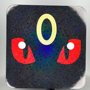 May include: Square-shaped sticker with a dark, glittery background and rounded corners. Features a yellow, oval-shaped design in the center, with red, angular shapes on either side, each with a white circle. The design resembles a stylized face.