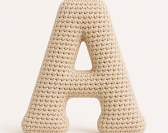 Puede incluir: Una letra A tridimensional hecha de hilo beige en un patrón de punto de ganchillo denso. La letra presenta una superficie uniforme y texturizada con una apariencia suave y redondeada, manteniéndose erguida con una estructura sólida.