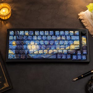 May include: A black mechanical keyboard with keycaps featuring a blue and yellow starry night design. The keyboard has a black knob and is surrounded by a candle, a book, and a pen.
