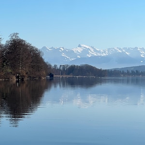 Könnte beinhalten: Eine ruhige Landschaft mit einem ruhigen See, der den klaren blauen Himmel und schneebedeckte Berge in der Ferne widerspiegelt. Kahle Bäume säumen das Ufer und bilden einen Kontrast zum ruhigen Wasser. Die Wasseroberfläche spiegelt Himmel und Berge wider.