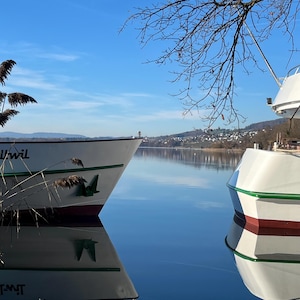 Könnte beinhalten: Zwei weiße Boote mit grünen und roten Verzierungen liegen auf einem ruhigen See vor Anker und spiegeln sich im Wasser. Die Boote stehen unter einem klaren blauen Himmel, mit ein paar kahlen Ästen in der oberen rechten Ecke. Das Wort "Uwil" ist sichtbar.