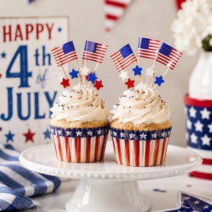 Könnte beinhalten: Zwei Cupcakes mit weißem Zuckerguss und rot-weiß-blauen Streuseln in patriotischen Förmchen. Jeder Cupcake ist mit kleinen amerikanischen Flaggen und Miniatursternen in Rot, Weiß und Blau verziert. Ein Schild trägt die Aufschrift Happy 4th of July.